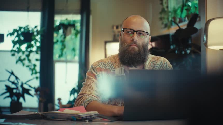 A thoughtful, bearded professional wearing glasses works from his plant-filled home office on a laptop