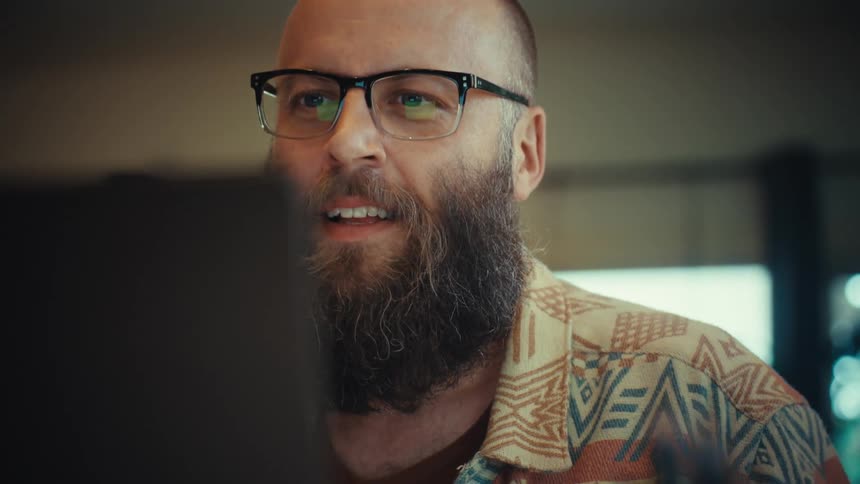 Close up of an expressive bearded man in glasses reacting with excitement, joy, and mischief to what he sees on his laptop