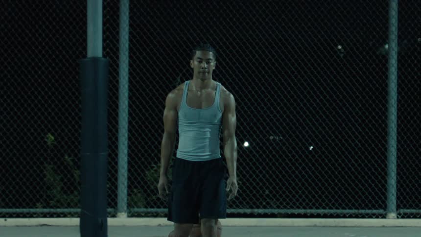 An athletic man dribbles a basketball and then walks towards the camera in slow motion on an outdoor court at night