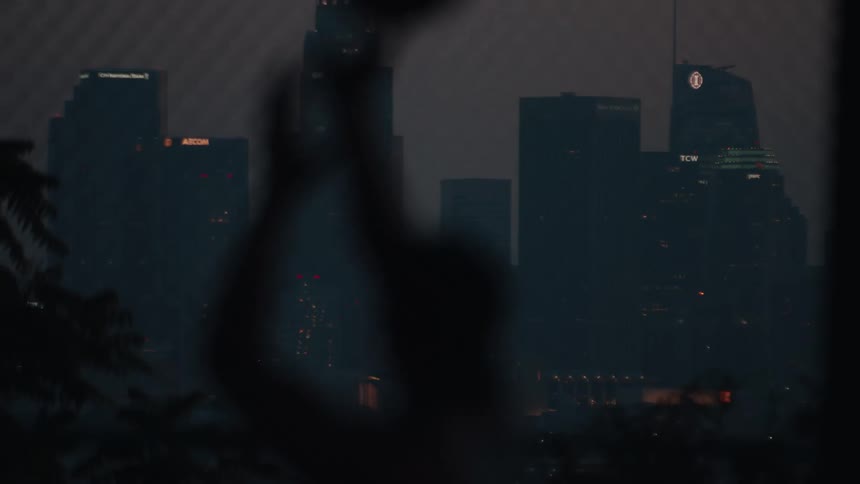 A blurry silhouette of a person raises their arms against the moody, illuminated downtown Los Angeles skyline at night