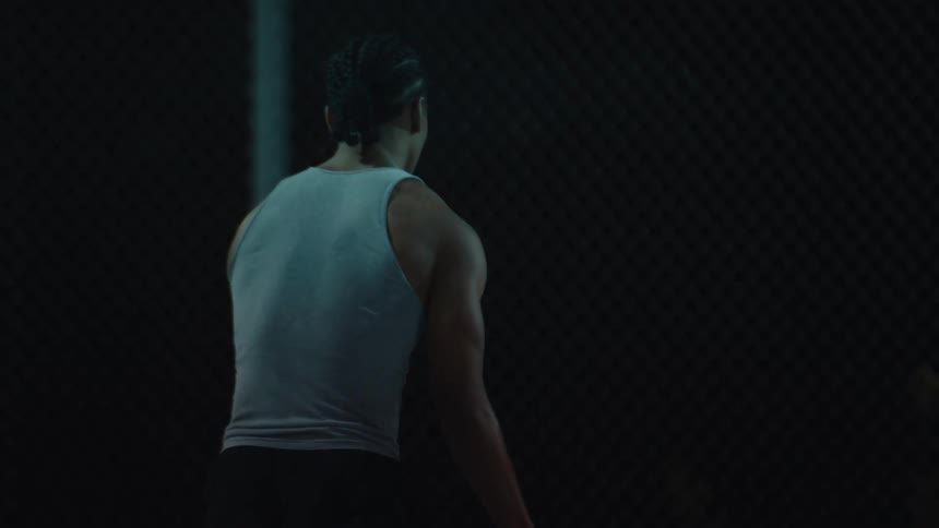 A muscular man with braided hair dribbles a basketball in slow motion on an outdoor court at night