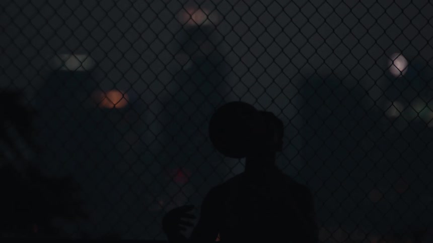 A silhouetted basketball player spins a basketball on their finger in slow motion against a chain-link fence with out-of-focus city lights at night
