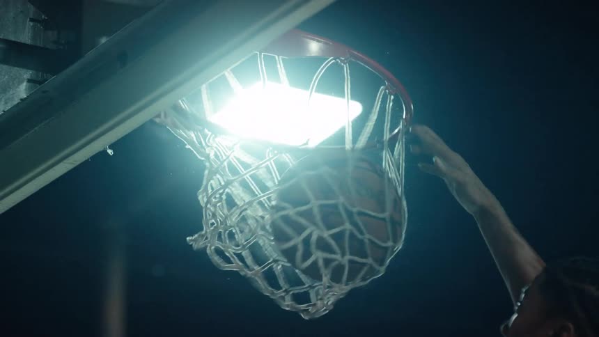 A basketball player slams a powerful dunk in dramatic slow motion on an outdoor court at night