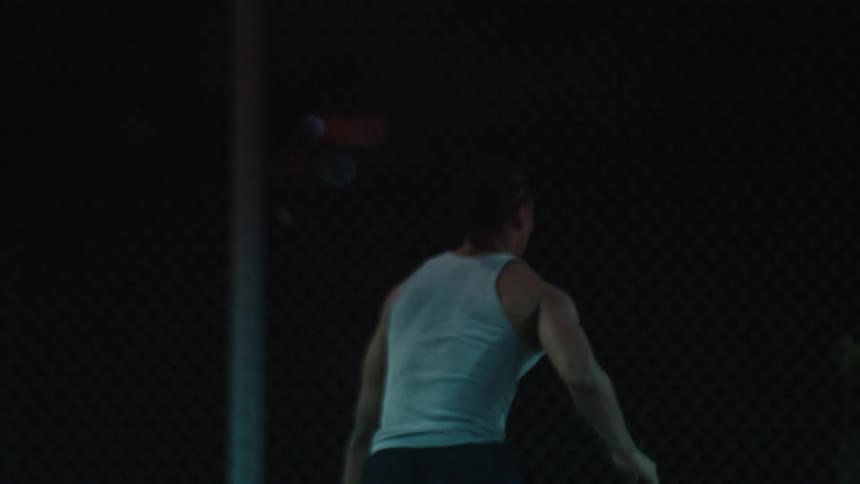 A muscular man seen from behind dribbles and takes a jump shot in slow motion on a dark city basketball court at night