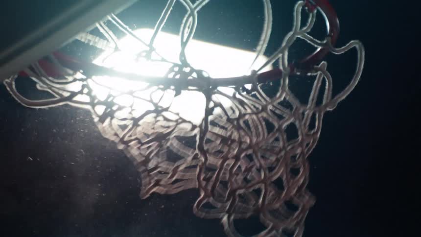 A basketball player scores a dramatic slam dunk in slow motion, seen from a low angle under a bright spotlight