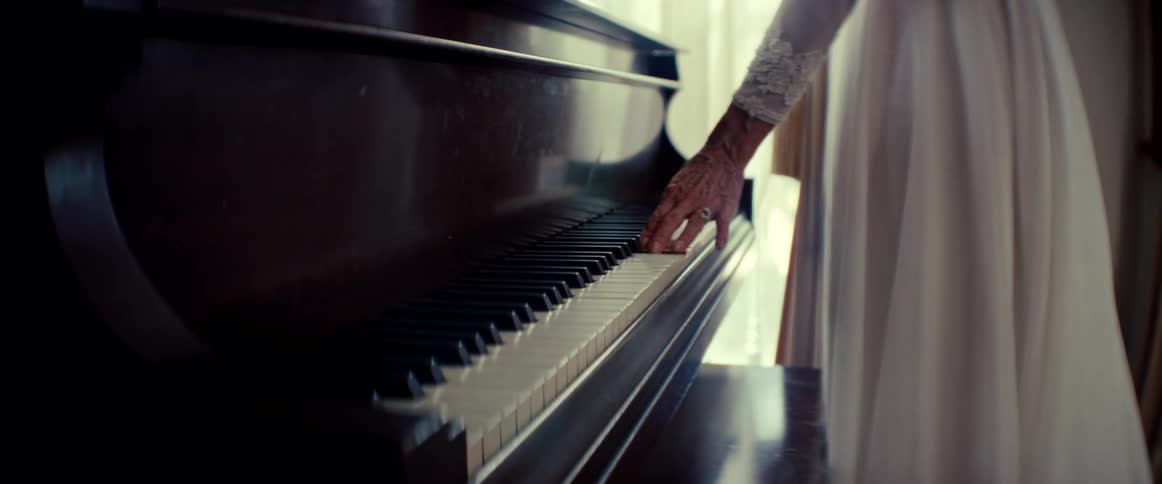 The wrinkled hand of an elderly woman in a white lace dress gently glides across the keys of a grand piano