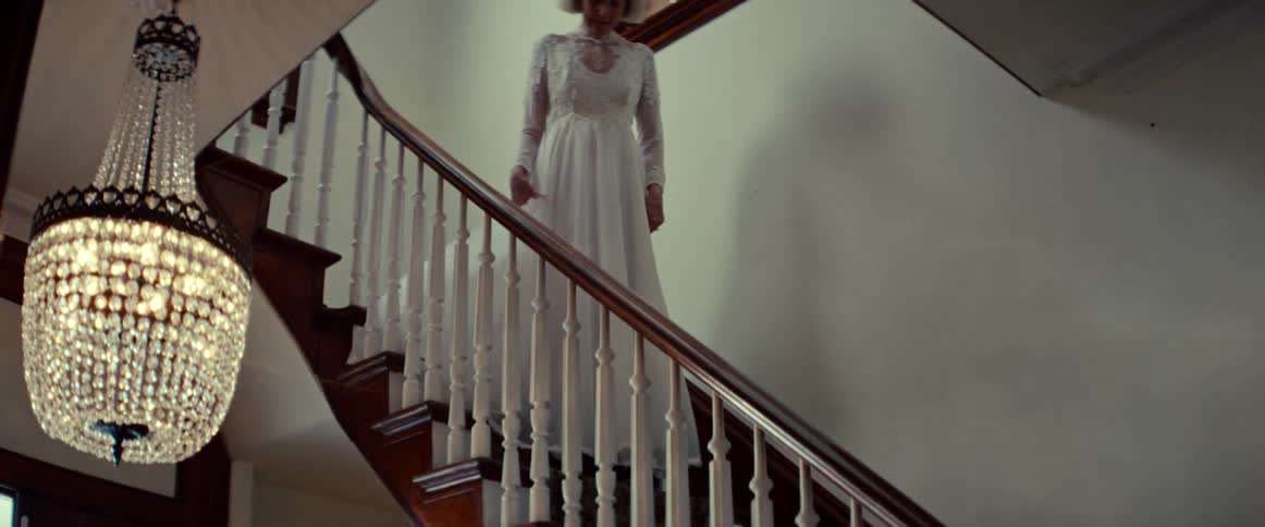 An elegant elderly woman in a vintage white wedding dress slowly descends a grand wooden staircase
