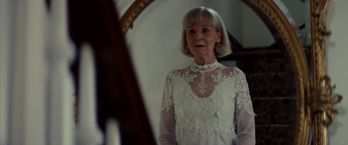 An elderly woman wearing a beautiful vintage wedding dress smiles at her reflection with a mix of joy and sadness