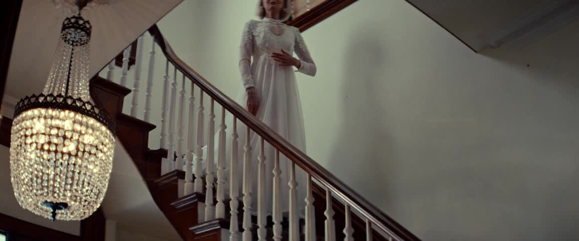 An elegant, wistful elderly woman in a vintage wedding dress stands at the top of a grand staircase