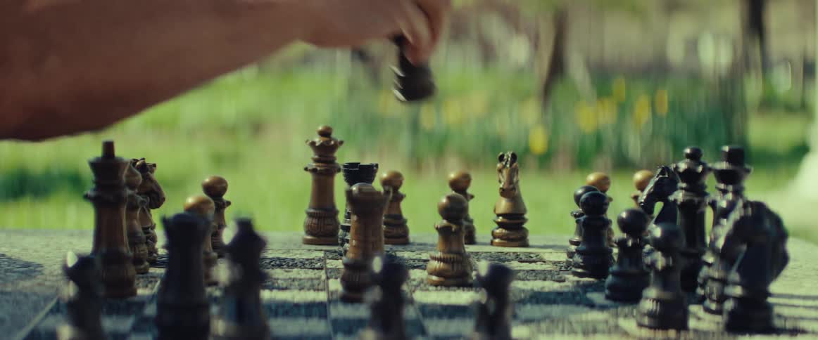 A person's hand makes a strategic move on a weathered chessboard during a game in a sunny, green garden