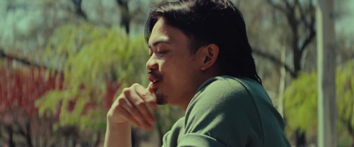 A happy young Asian man with a mullet enjoys a conversation in a park on a sunny day