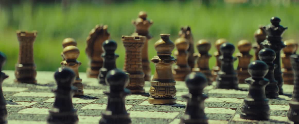 Close-up of a person's hand strategically moving a white bishop on a chessboard in a sunlit outdoor setting
