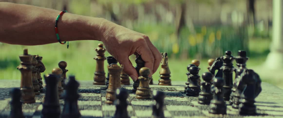 A person makes a strategic move with a black chess piece during a game on a stone table in a sunny park