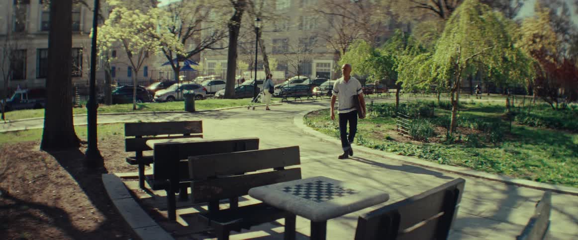 A man arrives at a sunny city park with his chessboard, looking for an opponent by the public chess tables
