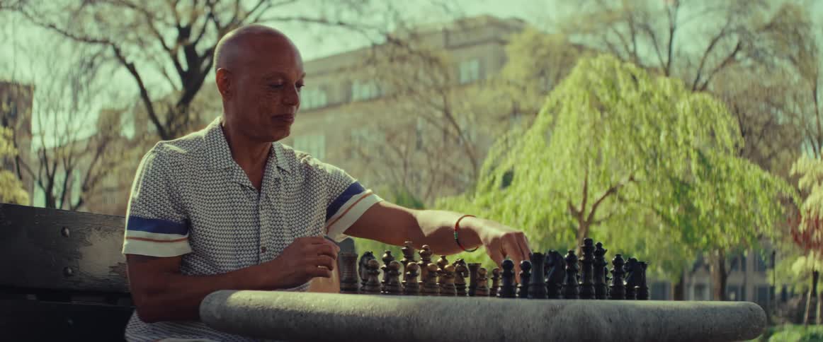 A thoughtful older man sits alone at a stone table in a park, contemplating and making a move in a game of chess