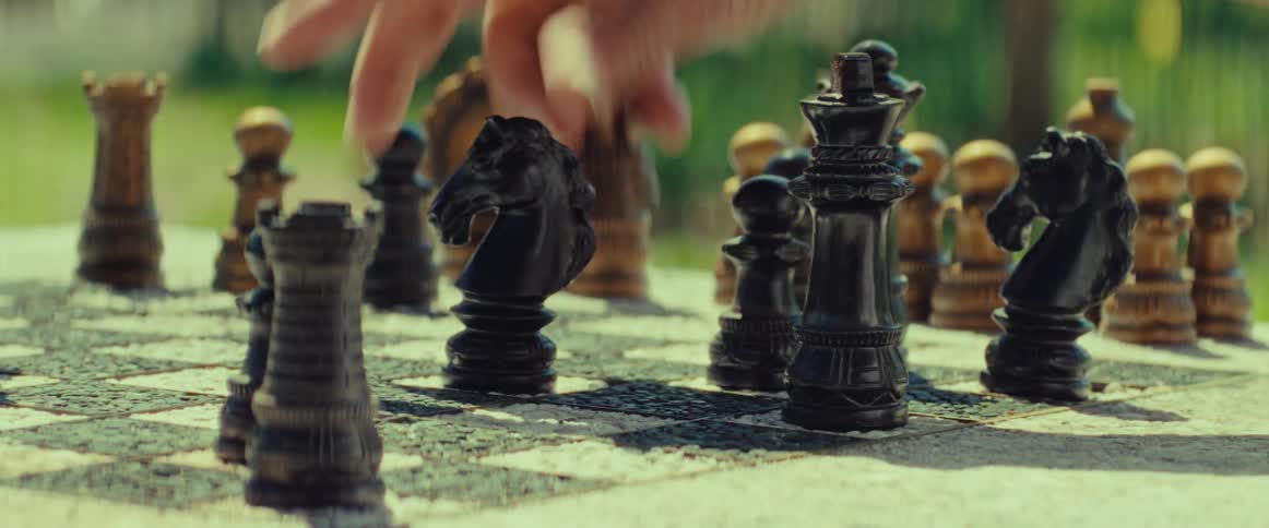 A person contemplates their next move before placing a black knight on an ornate chessboard outdoors