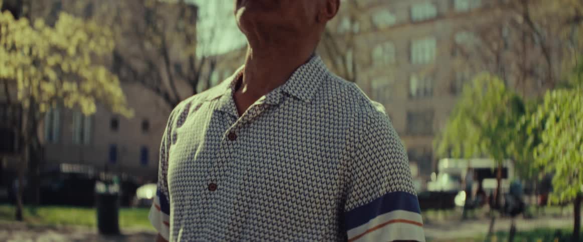 An older African American man holding a chessboard under his arm stands in a sunny city park and looks up thoughtfully