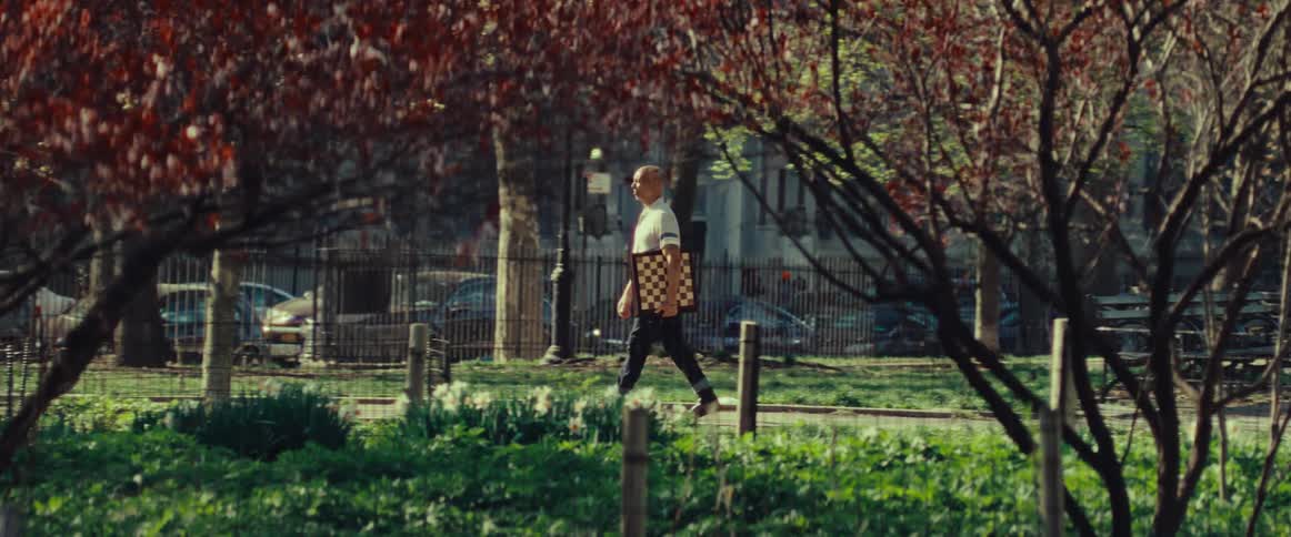 An elderly man carries a chessboard as he walks through a sunny city park in the spring