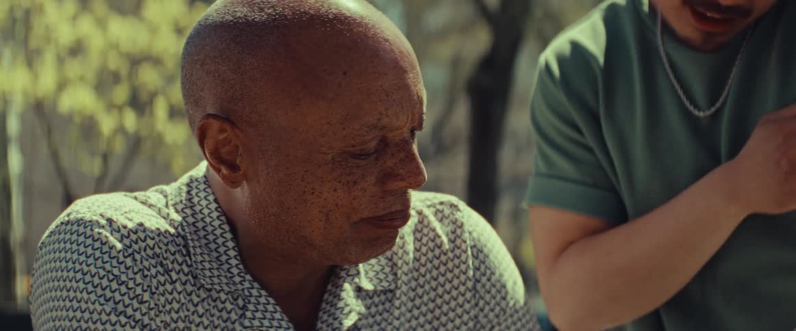 An older man and a younger man share a friendly conversation and a warm handshake while sitting in a sunny city park
