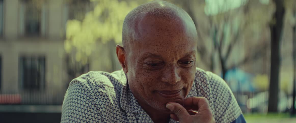 A thoughtful elderly Black man with freckles sits outdoors on a sunny day, a gentle, contemplative smile gracing his face