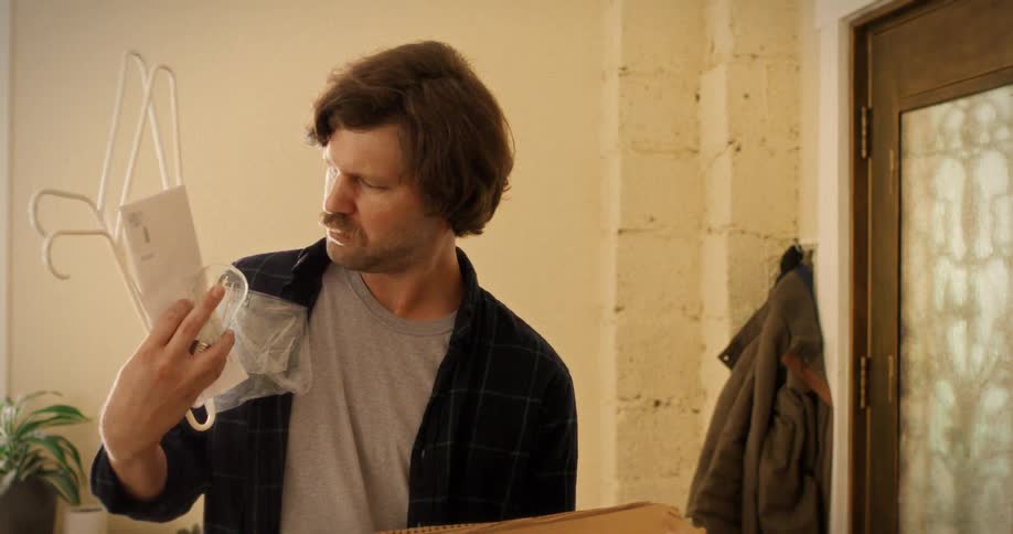 A man in a plaid shirt slowly unpacks a cardboard box and looks confused at the contents he pulls out