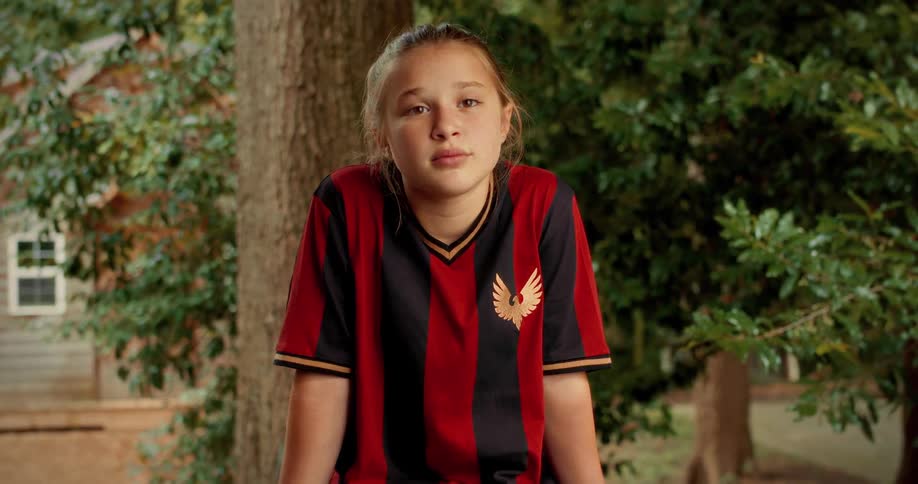 A confident young girl in a soccer jersey looks thoughtfully at the camera before giving a warm smile while sitting outdoors