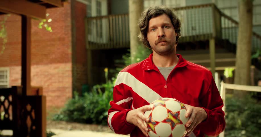 A mustached man in a retro tracksuit holds a soccer ball and stares down the camera in a suburban setting