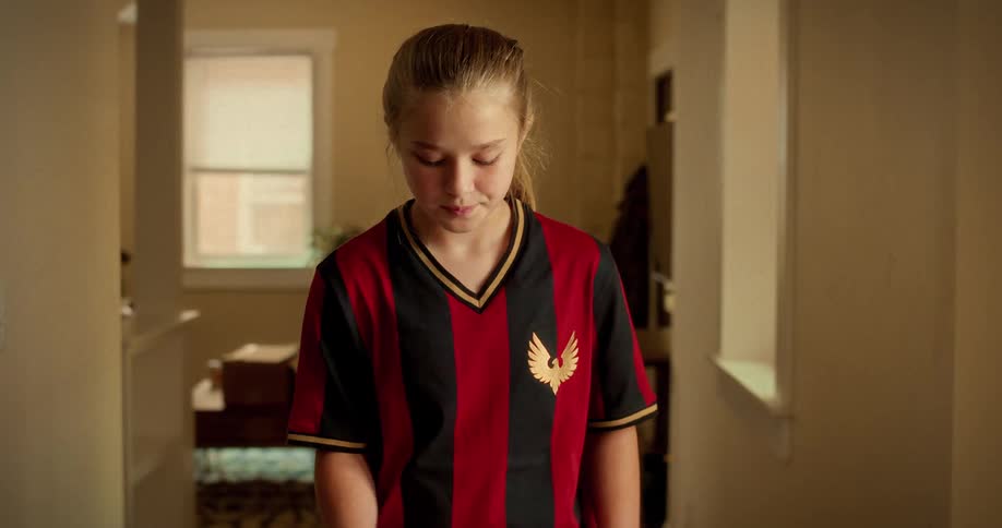 A young girl in a soccer jersey looks down with a sad expression before looking up at the camera in a sunlit hallway