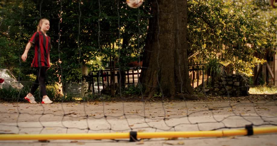 A young girl in a soccer jersey skillfully juggles a ball in slow motion and kicks it over a small net during practice