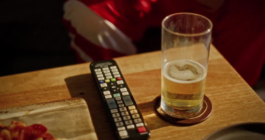 A close-up of a person in a red jersey placing a TV remote on a wooden table next to a glass of beer