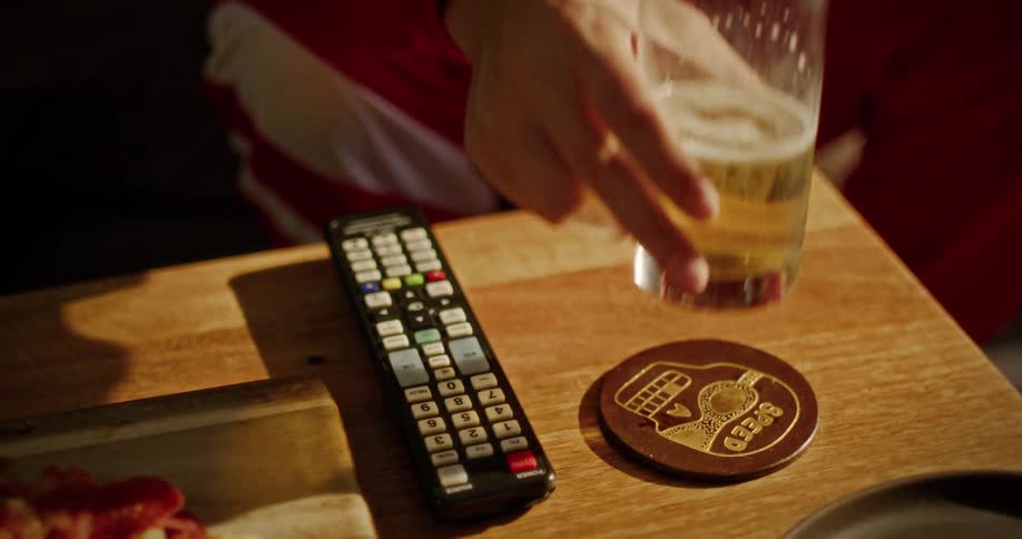 Slow motion close-up of a person taking a drink from a glass of beer on a table next to a remote control