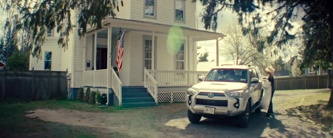 A woman packs a large white item into an SUV parked in the driveway of a classic American house