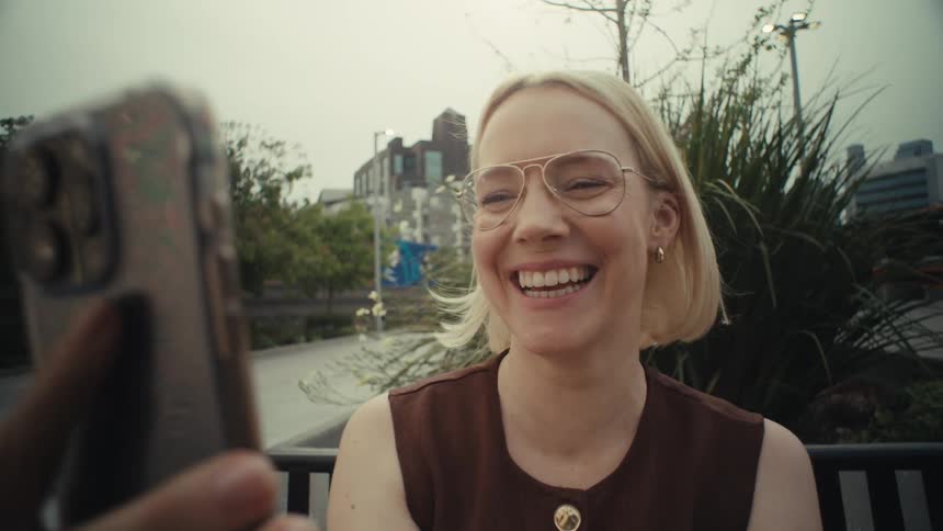 A close-up shot of a happy blonde woman with glasses laughing while looking at her smartphone in a city park
