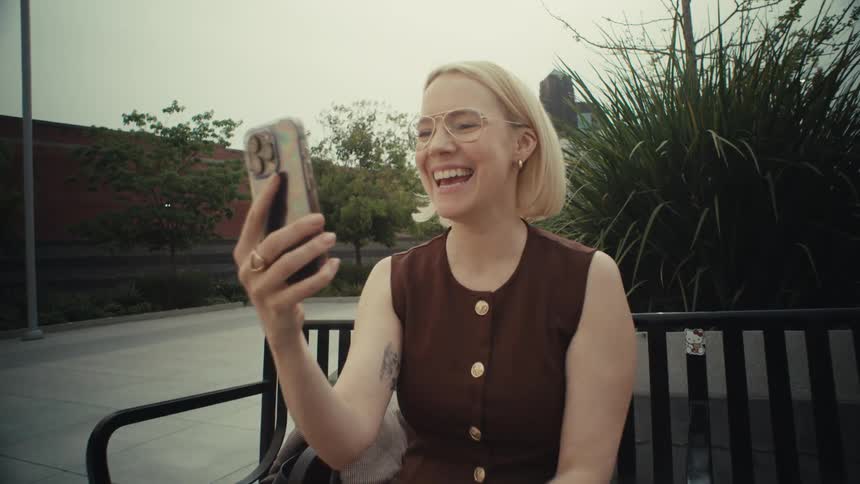 A happy woman laughs and waves during a video call on her phone while sitting on a bench in an urban park as the camera slowly zooms in