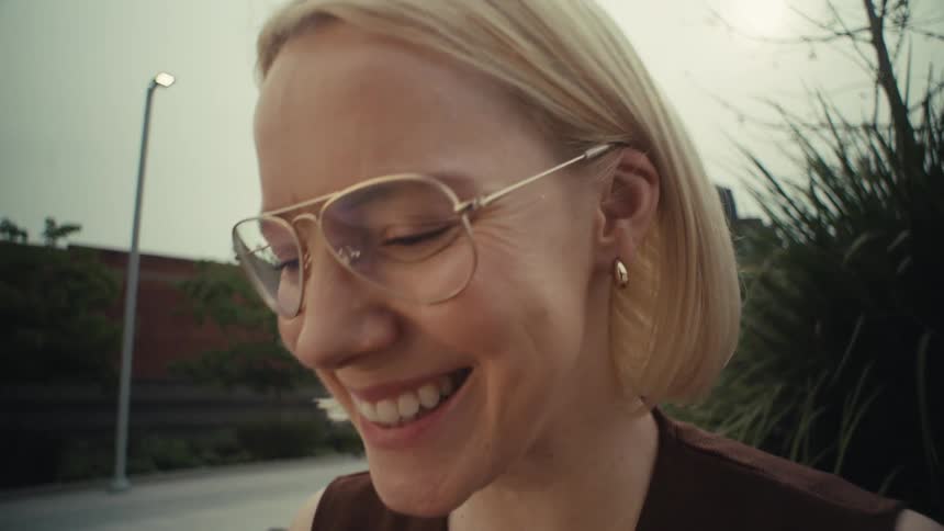 A joyful blonde woman with glasses smiles and laughs while looking at her smartphone as the camera slowly zooms out in an urban park