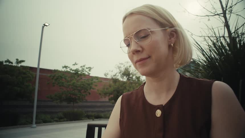 A slow-motion dolly shot pushes in on a stylish blonde woman with glasses using her smartphone on a city bench