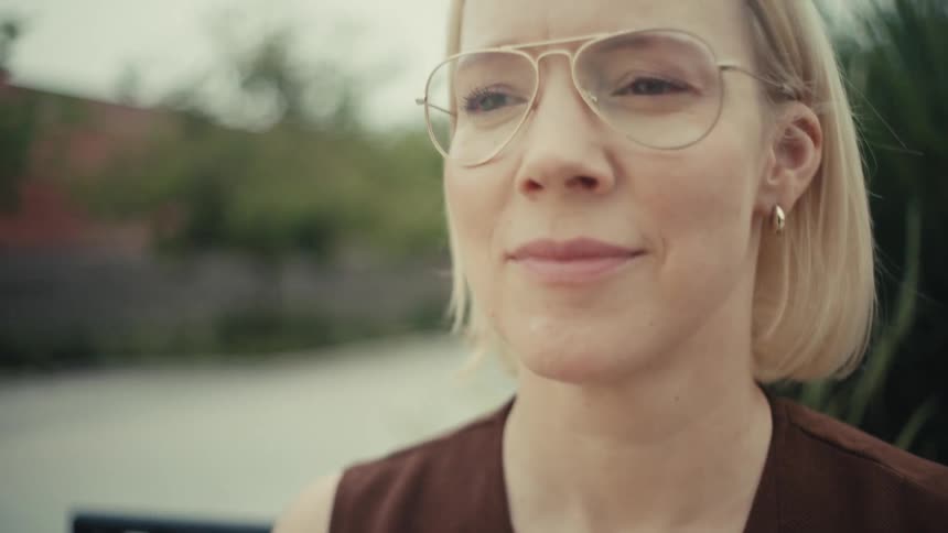 Close up on a stylish blonde woman with glasses using her smartphone while sitting on a park bench and smiling