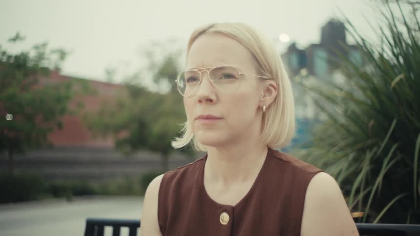 A pensive woman with blonde hair and glasses puts down her phone and gazes thoughtfully while sitting on a bench in a city park