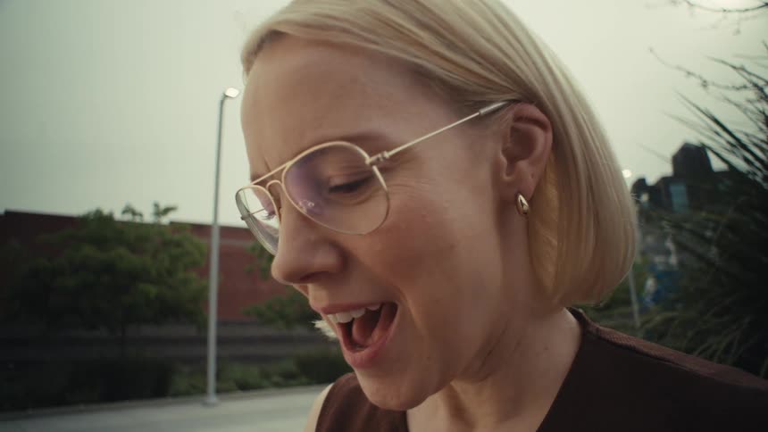 A joyous blonde woman in glasses laughs heartily while scrolling on her smartphone on a city park bench