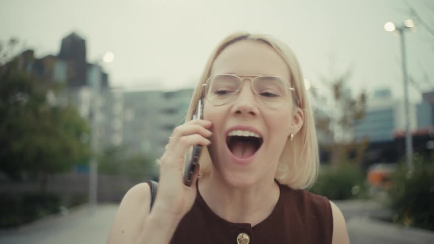 A stylish blonde businesswoman's expression shifts from neutral to ecstatic joy as she receives amazing news on a phone call while walking through the city