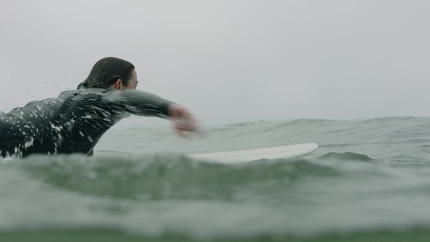 A female surfer in a wetsuit paddles on her surfboard in the ocean in slow motion on an overcast day