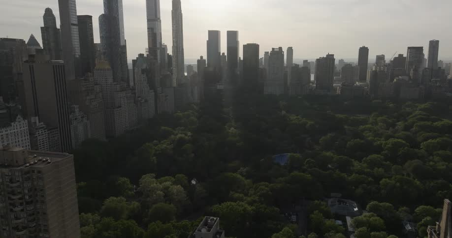 An aerial view slowly pushes forward over a lush Central Park surrounded by the iconic Manhattan skyline during a hazy sunrise
