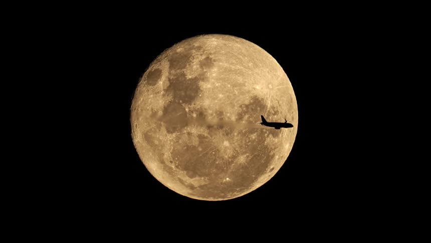 A silhouetted jet airplane with a contrail majestically crosses the face of a giant golden full moon at night