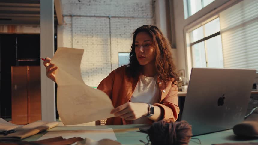 A thoughtful shoe designer inspects a hand-drawn sketch while working at her desk in an industrial loft