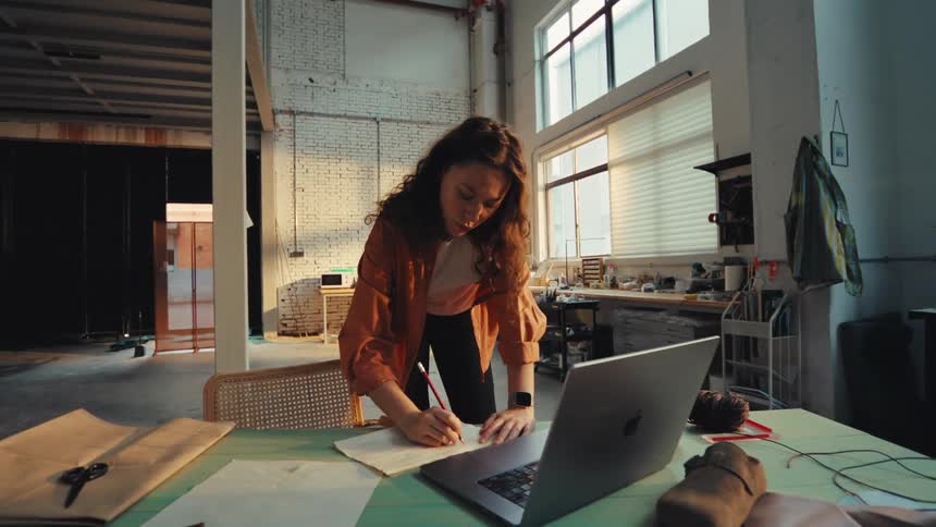 A focused young female artisan sketches a design on paper at her workbench in a sunlit industrial workshop