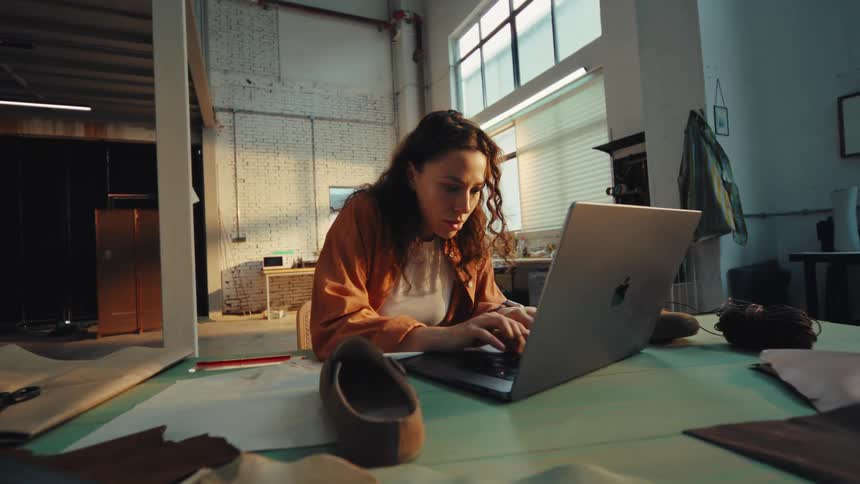 A diligent female artisan works on a new shoe design in her workshop, using a laptop for research and sketching ideas in her notebook