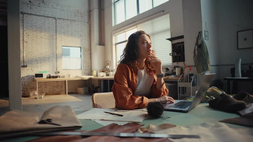 A thoughtful female artisan contemplates her next project while working on her laptop in a spacious, sunlit workshop