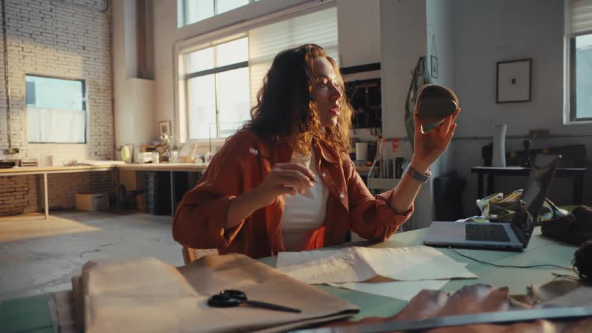 A creative female footwear designer sits at her desk, examining a shoe last and sketching a new design in her spacious, sunlit workshop