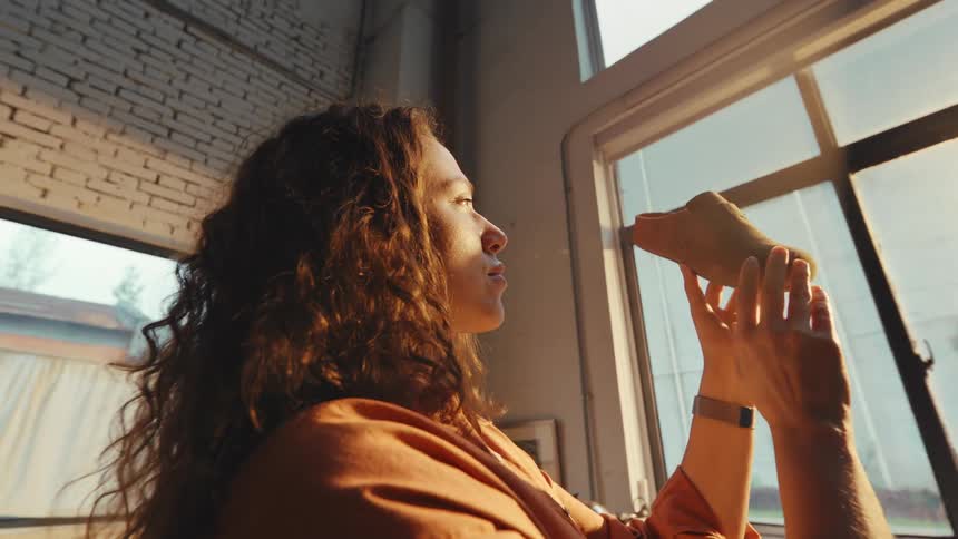 A female shoemaker with curly hair carefully inspects a handmade shoe in her sunlit workshop