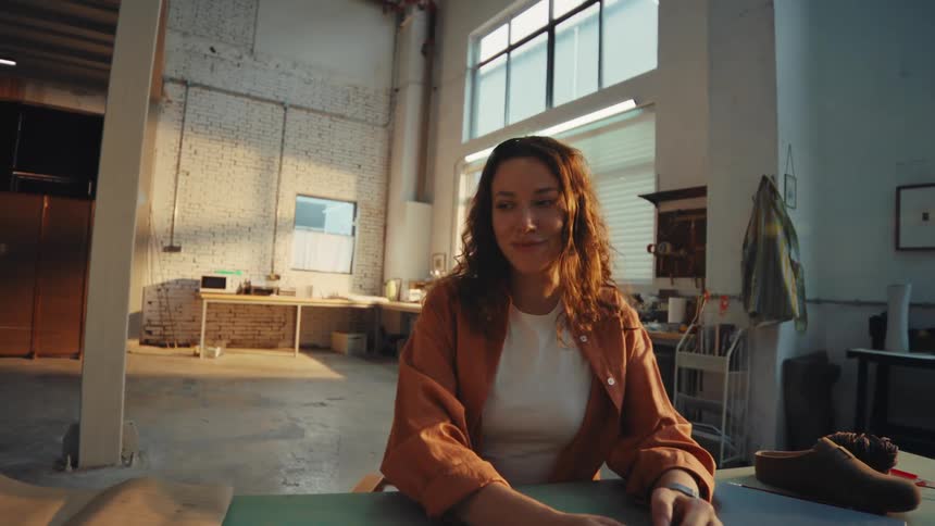 A female designer finishes typing on her laptop, closes it with a smile, and takes a moment of satisfaction in her spacious, sunlit workshop